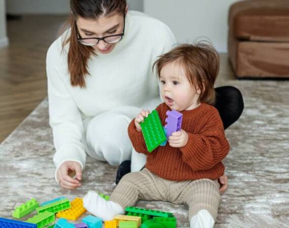 Babysitter and toddler playing with toy colored blocks at home. Happy family spend time together